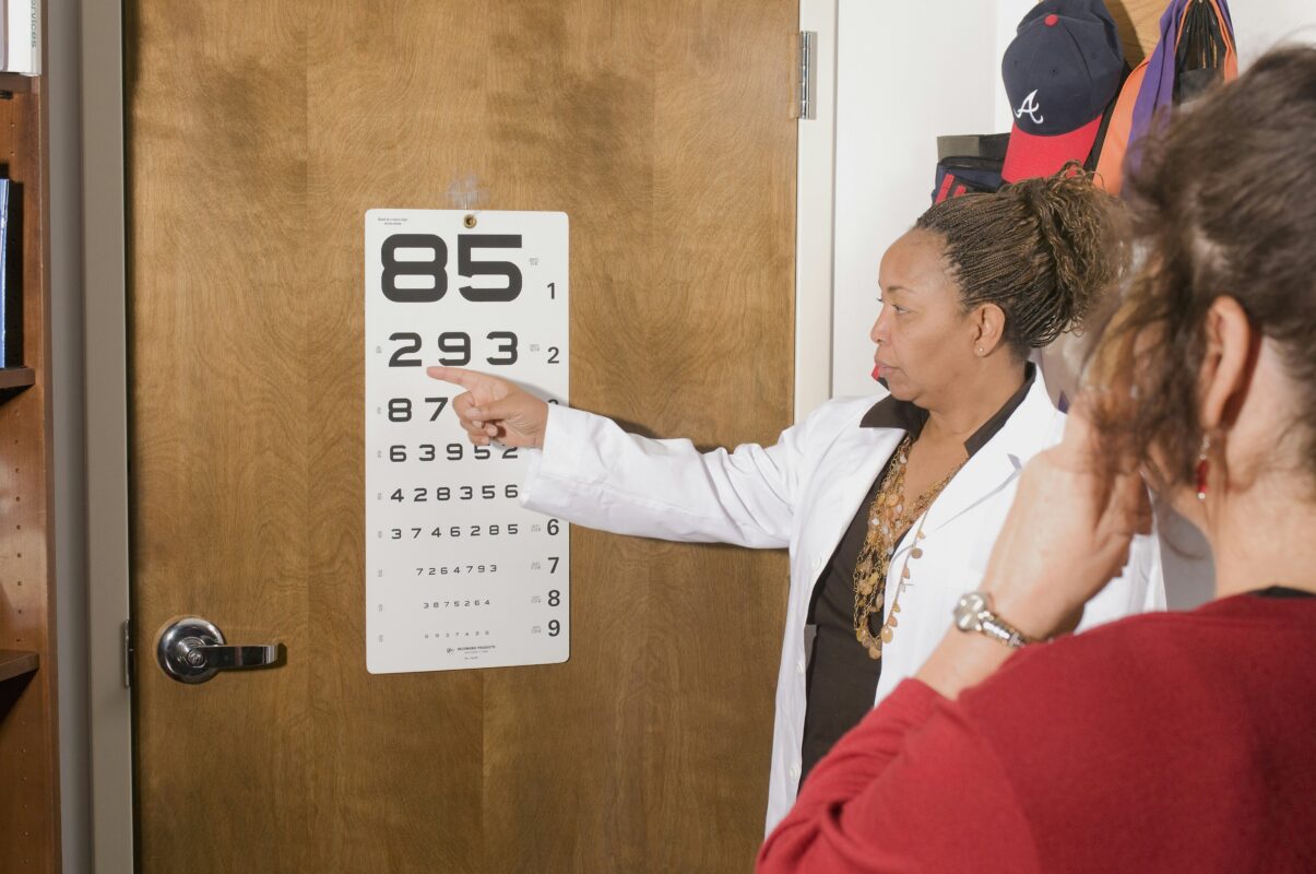 An eye doctor pointing to a wall-mounted number eye chart while conducting a vision screening test for a patient